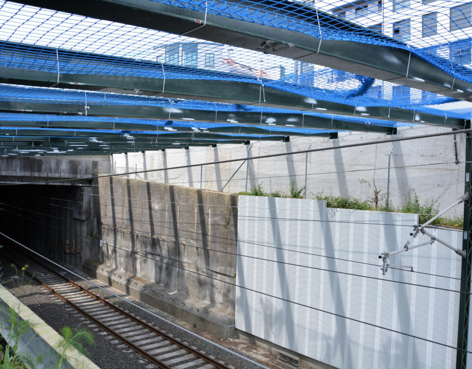Pared de hormigón junto a la vía del tren cubierta en parte por paneles acústicos bajo una estructura de vigas de acero